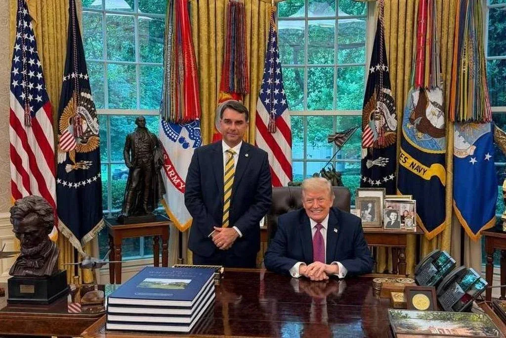 Dois homens posam no Salão Oval: um está sentado atrás da mesa presidencial e o outro em pé ao lado. Ao fundo, bandeiras dos Estados Unidos e da presidência, além de janelas com vista para árvores. O senador Flávio Bolsonaro (PL-RJ) posa para foto com Donald Trump na Casa Branca - 29.mai.26/@flaviobolsonaro no Instagram